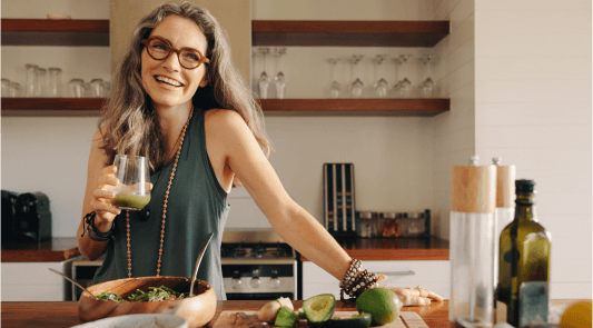 Woman smiling holding a juice cup in the kitchen with cutting board with cut avocados 